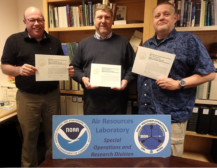 James S. Wood, Walter W. Schalk, and Ricky G. Lantrip standing in the SORD office holding their award certificates. The three are standing in front of a large panel displaying the NOAA and SORD logos.