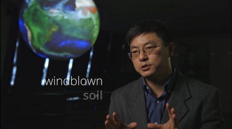 Dr. Tong with NOAA's Science on a Sphere behind him showing a video of windblown dust and the words "windblown soil" on the screen