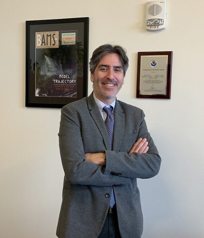 Dr. Stein standing in his office in front of a NOAA plaque and enlarged copy of an article that he authored