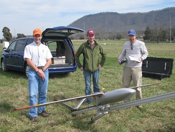Three people standing in a field behind the sUAS on its catapult.