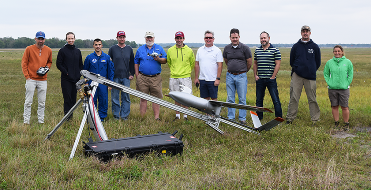 11 people standing in a field. Far left person holding a controller box, fifth from left holding an sUAS in one hand. Fixed-wing drone on a catapult with power case in front of the group.