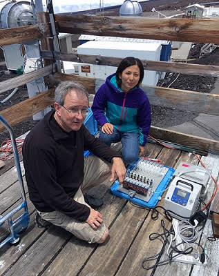 Two people kneeling on an elevated wooden deck attaching cabling to two instrument boxes.