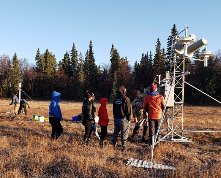 Two men with shovels in background at left. Seven people beside metal tower, one gesturing to/explaining meteorological instruments on the tower.