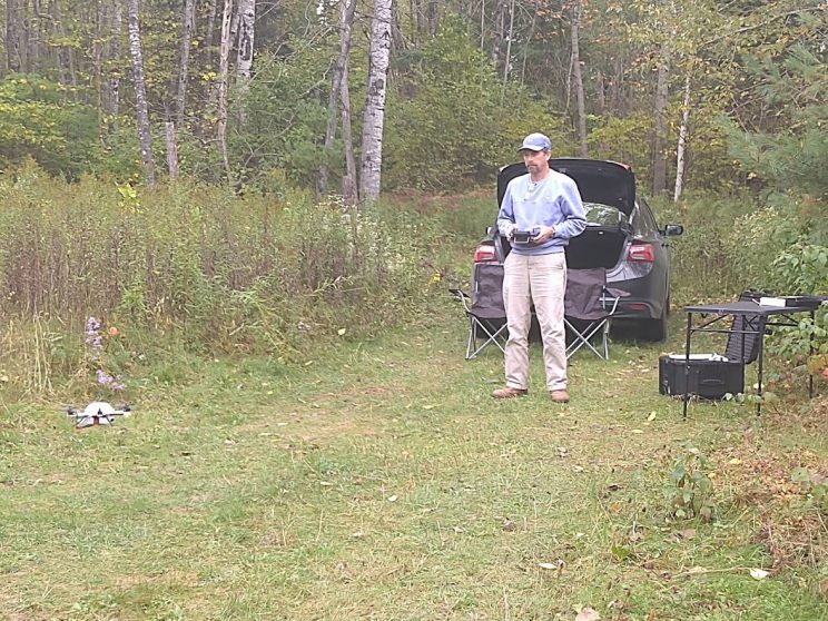 Small drone lifting off the ground in a small clearing at the edge of woods. A man stands a few feet behind it using the control box.