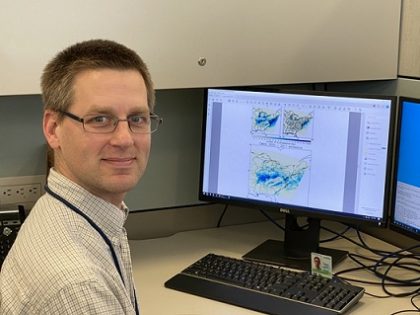 Dr. Loughner sitting at his computer, facing the camera. His monitor displays model results from the study: two small images above one large image, all color-coded maps of the Eastern US.