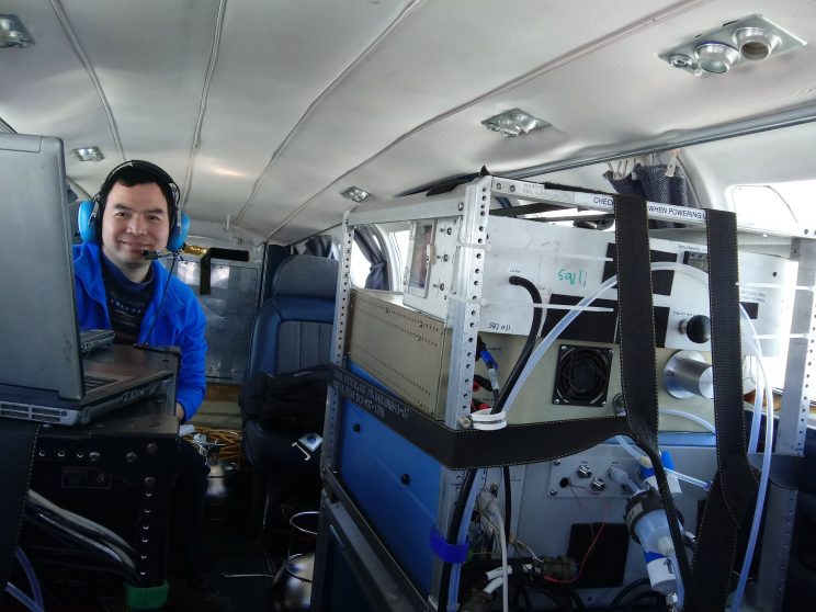 View from the cockpit looking back into the plane. On the left, a man with a headset on is behind two open laptops on top of a metal shelving unit containing what looks like metal boxes with tubes and cables. On the right, the back of a metal rack containing similar, but larger metal instrument boxes is strapped around the sides and top to anchor it in place.