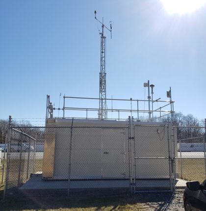 Inside a chain link fence is a metal trailer topped with a two-tiered metal railing. Various rectangular and cylindrical shaped devices are attached to metal poles on the railing and a scaffold.