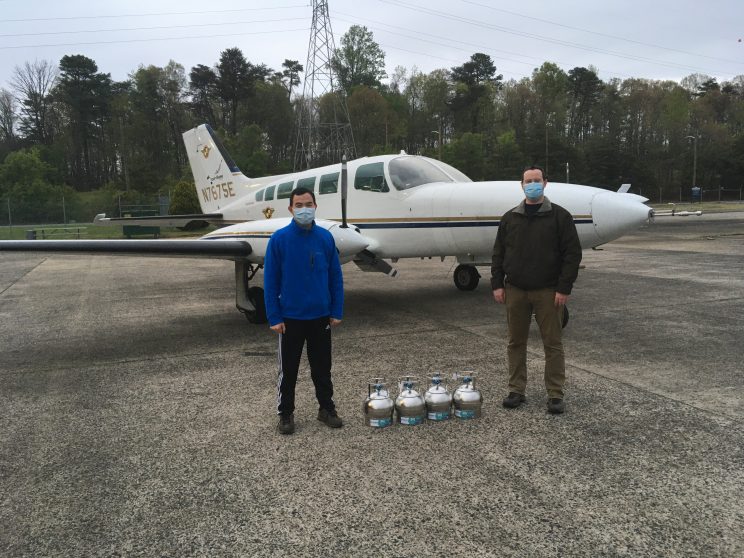 Two men wearing medical masks stand in front of a small aircraft. Four small metal cylinders are on the ground between them.