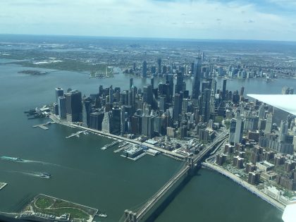 View out the window of the research aircraft, wing visible at right. Plane is above the Brooklyn Bridge and the buildings of Southern Manhattan are visible, surrounded on three sides by calm water.