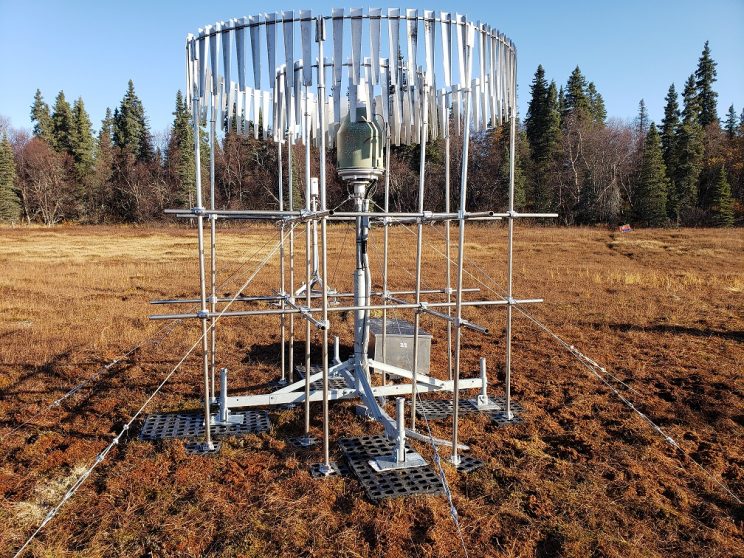A metal grid structure surrounds a thin metal post, on top of which sits what looks like a metal bucket with a tapered top. Long, thin strips of metal hang in two circles, one inside the other, at the top of the structure, which is tethered to the ground by metal cables.