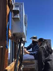 Metal box containing instrumentation on the top of mauna loa as two people service it.