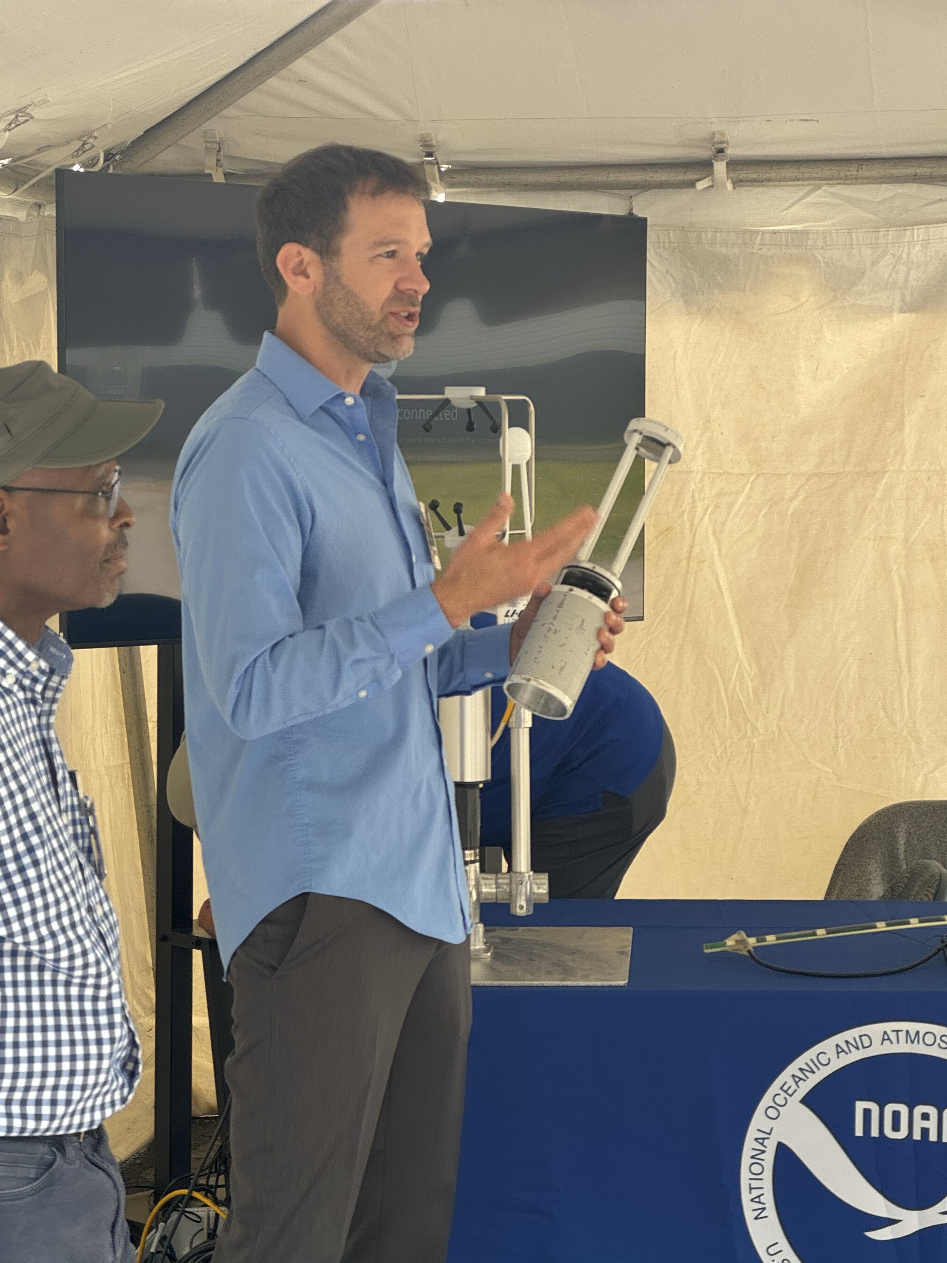 Two men standing, the one on the right holds a sensor and gestures as he speaks.