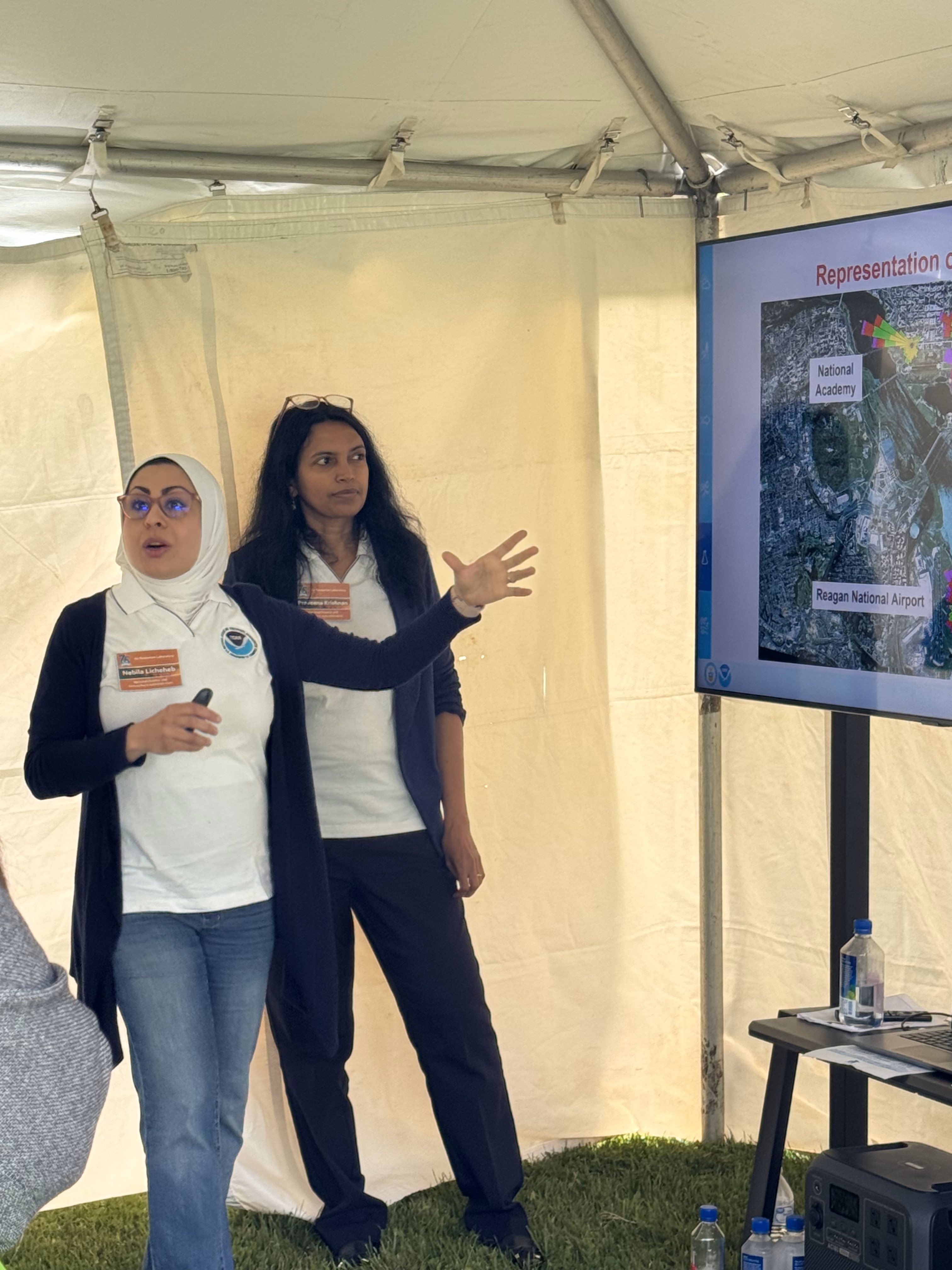 Two women standing inside a tent, presenting material that appears on a screen beside them