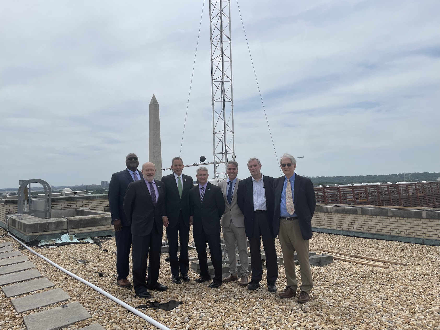 Group of 7 men in suits standing on a flat rooftop with a metal frame tower on the roof behind them and the Washington Monument in the background.