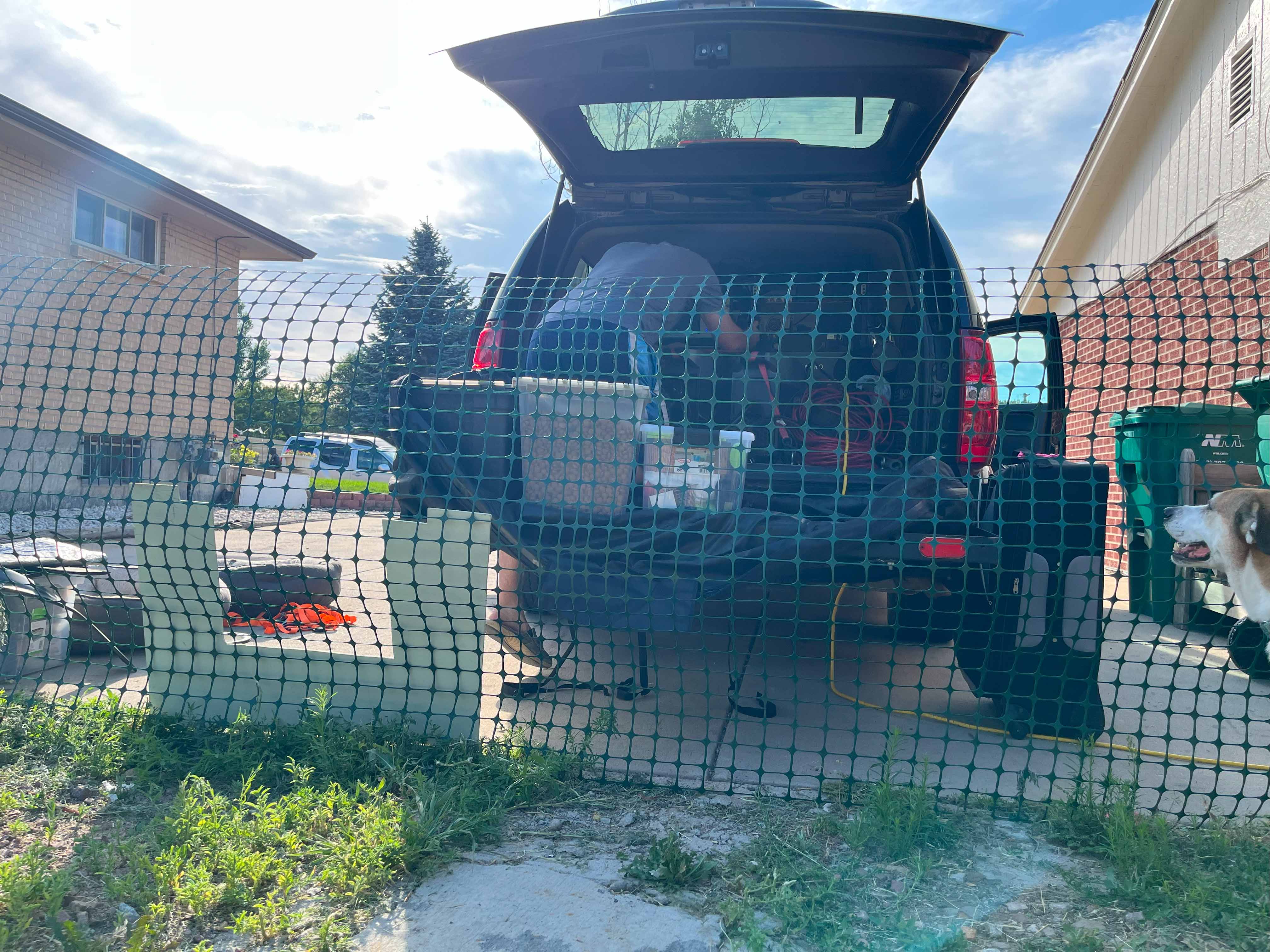 photo of the back of an SUV with a man loading items into the back, there is a fence behind the car, supplies waiting to be loaded and a dog in the corner watching the loading process.