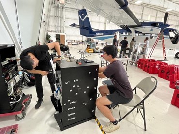 two men working on the instruments in a rack on the floor of a hanger. A propeller plane is in the background.