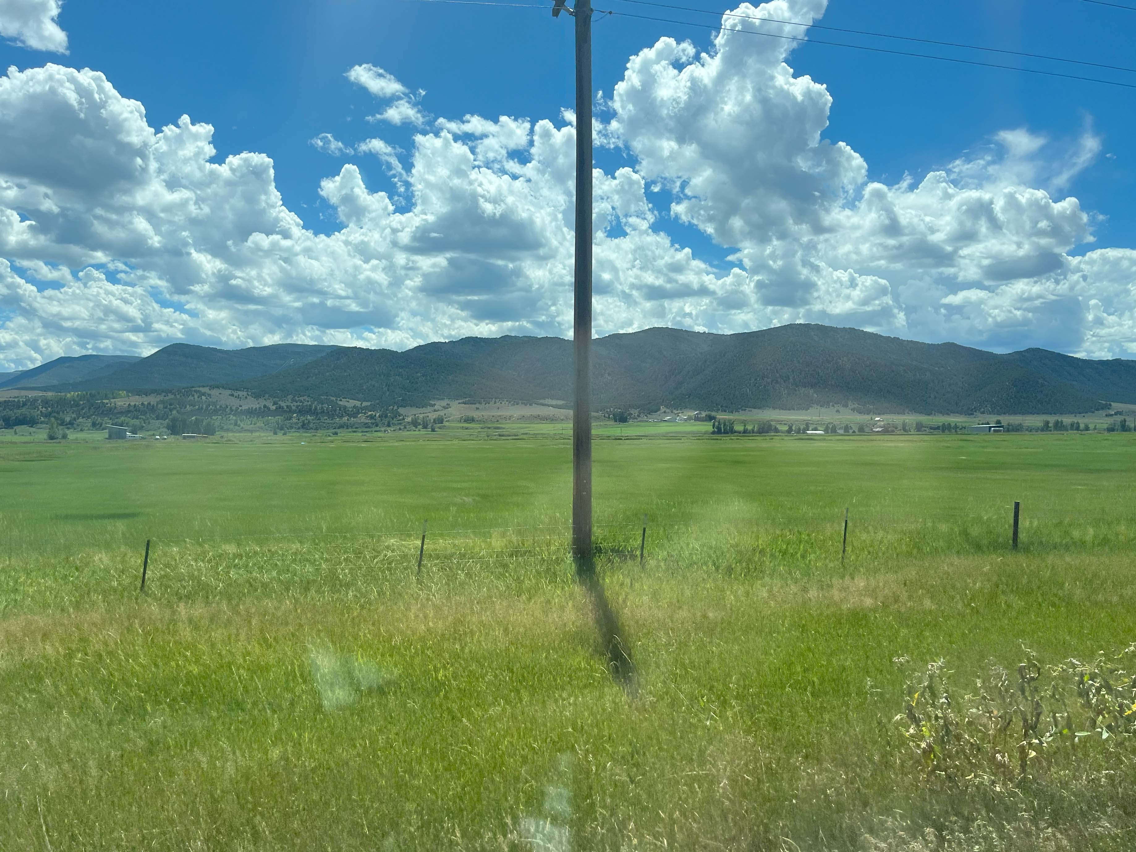 Field on a bright sunny day with mountains in the background and clouds in the sky. A telephone pole is in the middle of the image.